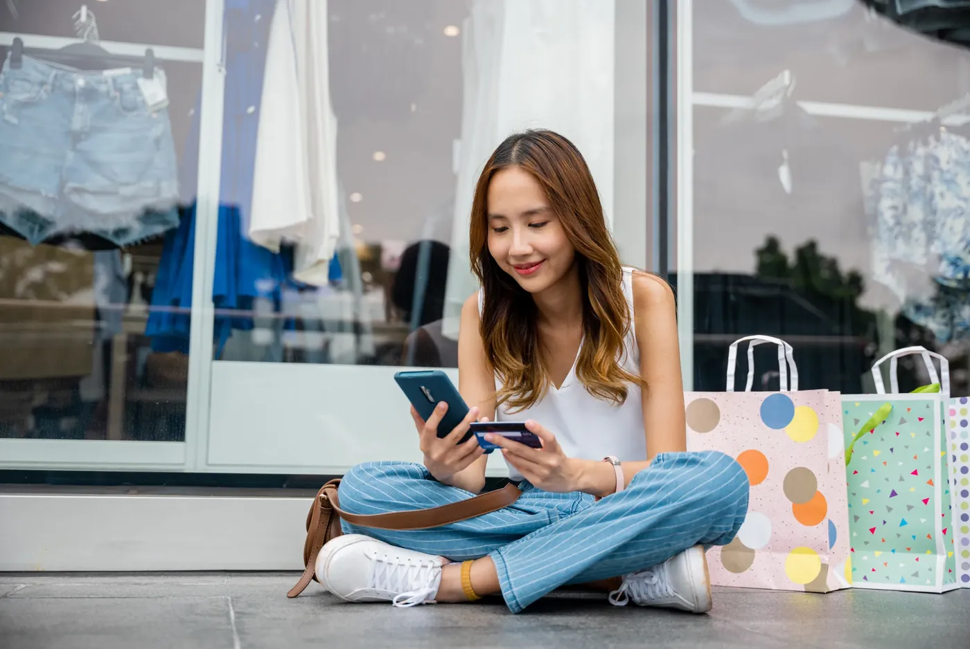 A young woman sitting outside a clothing store, browsing her phone with a payment card in her hand