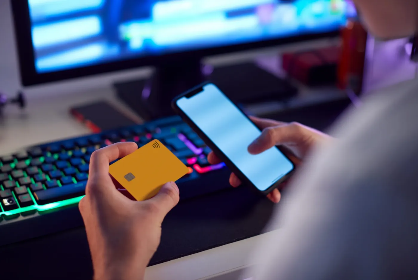 A person holding a payment card and phone late at night in front of a glowing screen